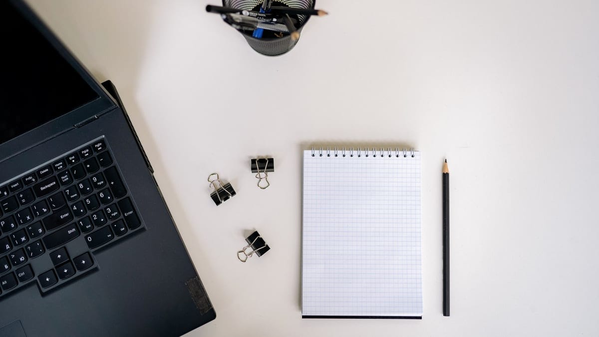 Top view of an office setup with a laptop, notepad, pencils, and clips on a white desk.