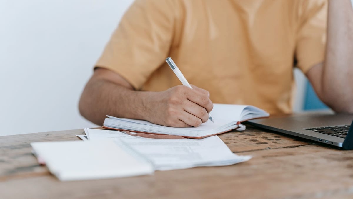 Crop anonymous male in casual wear working on netbook and taking notes in notepad while sitting at desk
