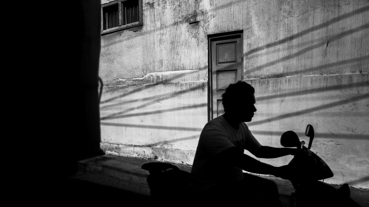 A stark black and white silhouette of a person riding a scooter against an urban wall with window shadows.