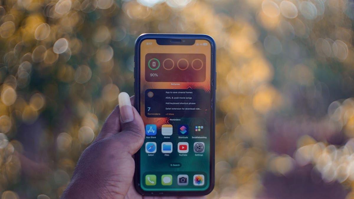 A hand holding a smartphone showing home screen apps, outdoors with bokeh background.