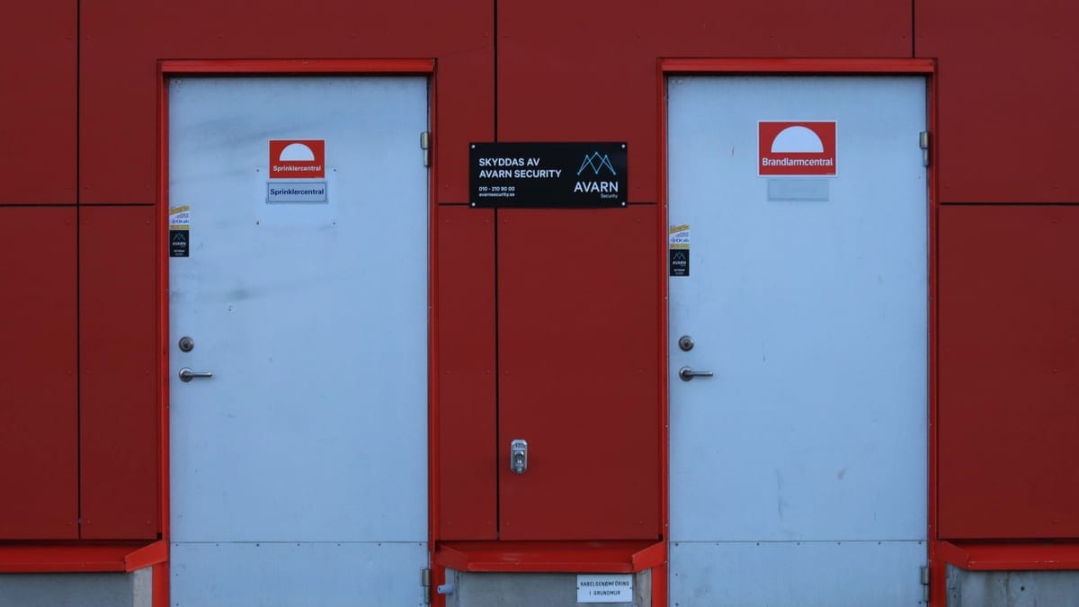 Two white doors against a red wall featuring security signs and logos.